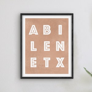 May include: A framed print with a brown background and white text that reads "ABILENE TX". The print is hanging on a white wall with a clock and a lamp on a white table.