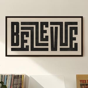 May include: A black and white print of the word "BELIEVE" in a geometric font, framed in black and hanging on a white wall. The print is above a record player, a stack of vinyl records, and a record album with the text "BEST OF THE BEACH BOYS" on the cover.