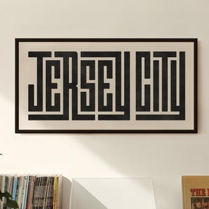May include: A black and white framed print with the text "JERSEY CITY" in a bold, geometric font. The print is hanging on a white wall above a wooden shelf with a record player, a stack of vinyl records, and a record album.