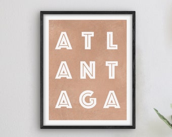 Könnte beinhalten: Ein gerahmter Druck mit kupferfarbenem Hintergrund und weißem Text, der "ATL ANTA GA" lautet. Der Druck hängt an einer weißen Wand neben einer Uhr und einer Lampe.