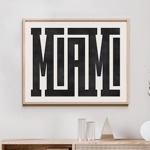 K&ouml;nnte beinhalten: Ein Schwarz-Wei&szlig;-Typografie-Druck mit dem Wort "MIAMI" in fetten Blockbuchstaben. Der Druck ist in einem hellen Holzrahmen gerahmt und h&auml;ngt an einer wei&szlig;en Wand &uuml;ber einem Holzregal.