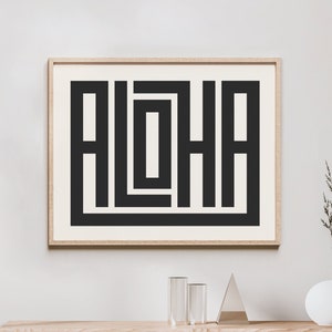 K&ouml;nnte beinhalten: Ein gerahmter Druck mit einem geometrischen Design in Schwarzwei&szlig; des Wortes "ALOHA". Der Druck h&auml;ngt an einer wei&szlig;en Wand &uuml;ber einem Holzregal mit einer Vase mit getrockneten Blumen.