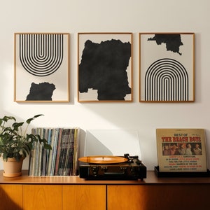 Puede incluir: Tres impresiones de arte abstracto en blanco y negro con formas geométricas cuelgan de una pared blanca sobre un armario de madera con un tocadiscos y una pila de discos de vinilo. Una planta en maceta se encuentra sobre el armario.
