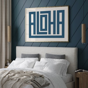 May include: A bedroom with a white headboard and a framed print with the word "ALOHA" in blue letters on a white background. The print is hanging on a dark blue wall with white diagonal stripes. The bed has a white duvet and a beige throw blanket.