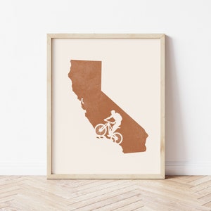 May include: A framed print of the state of California in a burnt orange color with a white silhouette of a person riding a mountain bike.