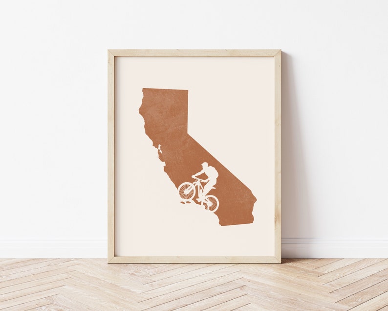 May include: A framed print of the state of California in a burnt orange color with a white silhouette of a person riding a mountain bike.