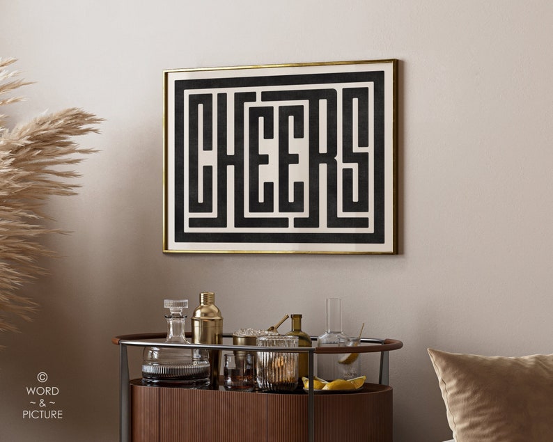 May include: Black and white typography art print with the word "CHEERS" in a geometric design. The print is framed in a gold frame and hanging on a light beige wall.