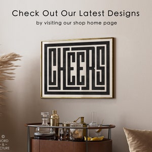May include: Black and white geometric print with the word "CHEERS" in a maze-like design. The print is framed in a gold frame and hanging on a beige wall.