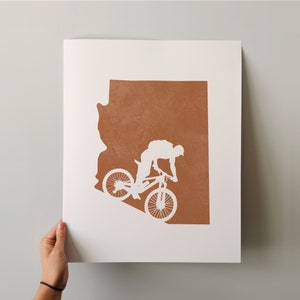 May include: A white silhouette of a person riding a mountain bike on a brown outline of the state of Arizona.