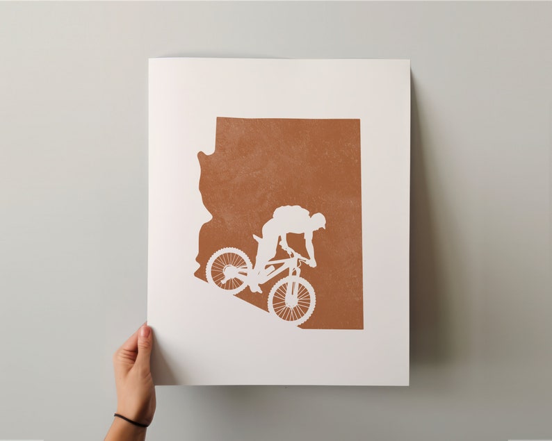 May include: A white silhouette of a person riding a mountain bike on a brown outline of the state of Arizona.