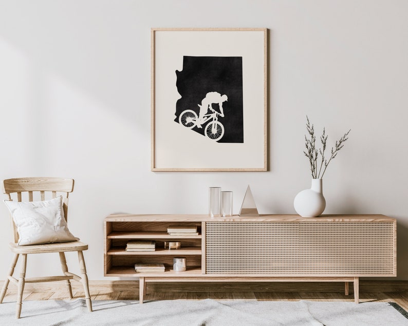May include: A black and white print of the state of Arizona with a silhouette of a person riding a mountain bike. The print is framed and hanging on a white wall above a wooden cabinet.
