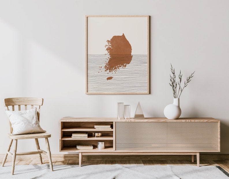 May include: A light wood media console with a woven rattan front and open shelves. A white vase with dried branches sits on top of the console. A framed print of a brown silhouette of South Korea with white lines is on the wall behind the console.