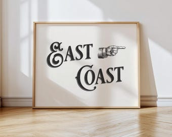 Pode incluir: Uma impressão emoldurada com as palavras "East Coast" em uma fonte decorativa. Uma mão apontando para a direita está ao lado da palavra "Coast". A impressão está em uma moldura dourada e está encostada em uma parede branca em um piso de madeira.