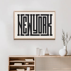 May include: A framed print with the word "NEW YORK" in a bold, black, geometric font. The print is hanging on a white wall above a wooden cabinet with shelves and a woven rattan front.