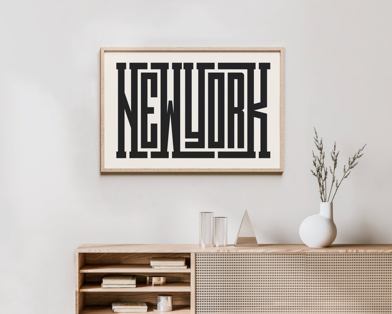May include: A framed print with the word "NEW YORK" in a bold, black, geometric font. The print is hanging on a white wall above a wooden cabinet with shelves and a woven rattan front.