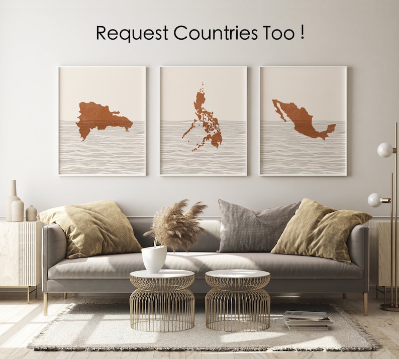 May include: Three framed prints of country outlines in a copper color on a white background. The outlines are of the Dominican Republic, the Philippines, and Mexico. The prints are hanging on a white wall above a gray sofa with pillows and a vase of dried flowers.