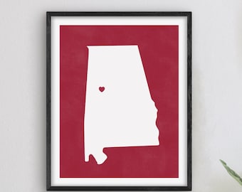 Op de afbeelding: Een witte omtrek van de staat Alabama op een rode achtergrond met een klein rood hart in het midden van de staat.