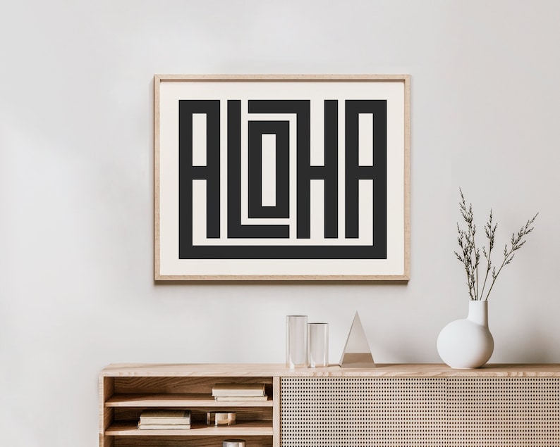 K&ouml;nnte beinhalten: Ein gerahmter Druck mit einem geometrischen Design in Schwarzwei&szlig; des Wortes "ALOHA". Der Druck h&auml;ngt an einer wei&szlig;en Wand &uuml;ber einem Holzregal mit einer Vase mit getrockneten Blumen.