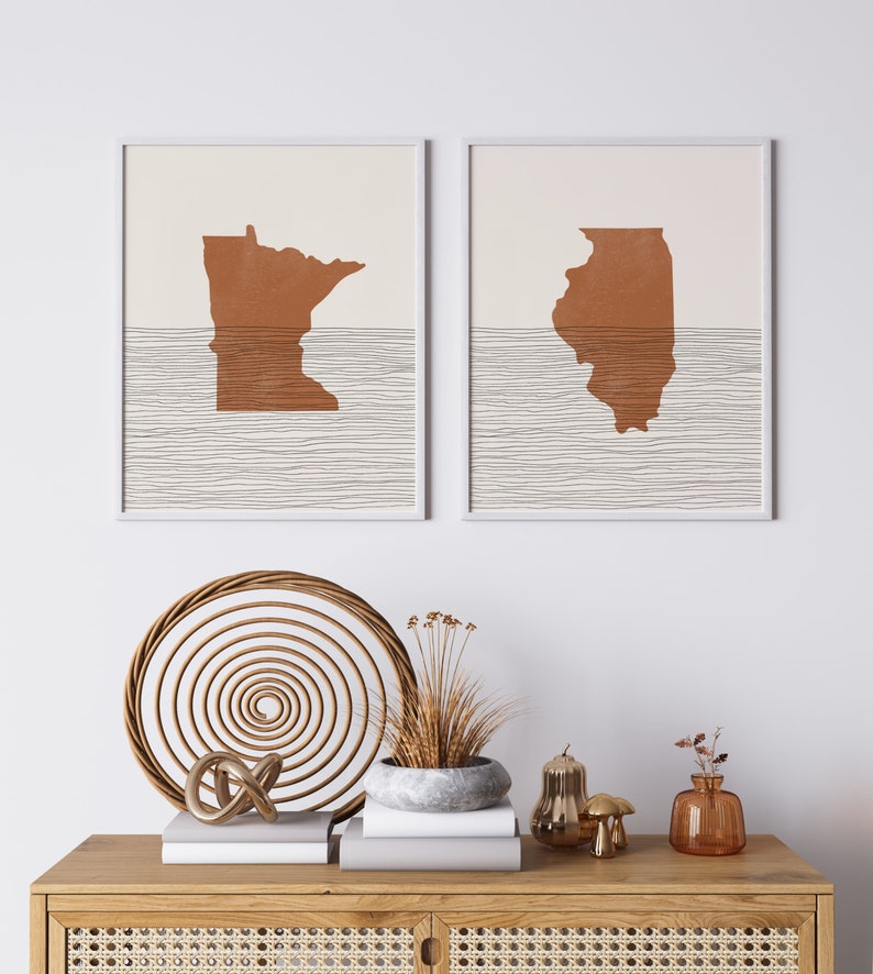 May include: Two framed prints of the states of Minnesota and Illinois, with a brown silhouette of each state on a white background with brown lines. The prints are hanging on a white wall above a wooden dresser with a wicker basket, a potted plant, and decorative objects.