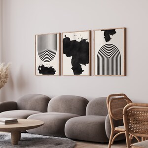 May include: Three black and white abstract art prints with geometric shapes and brushstrokes in wooden frames hanging on a white wall above a gray sofa and wicker chairs.