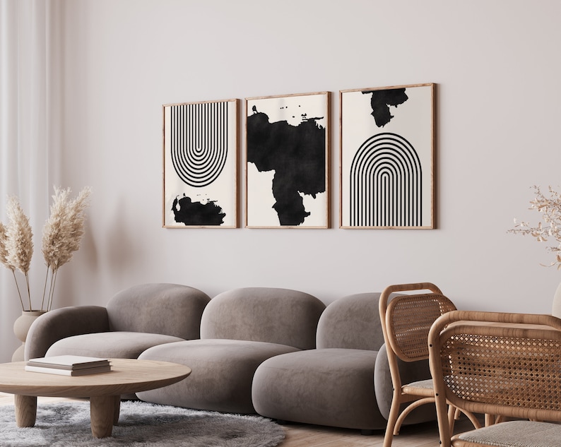 May include: Three black and white abstract art prints with geometric shapes and brushstrokes in wooden frames hanging on a white wall above a gray sofa and wicker chairs.