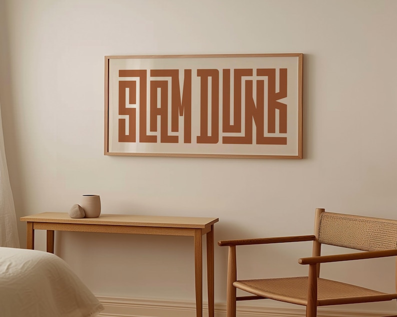 May include: A framed print with the text "SLAM DUNK" in a brown color. The print is hanging on a white wall above a wooden table and chair.