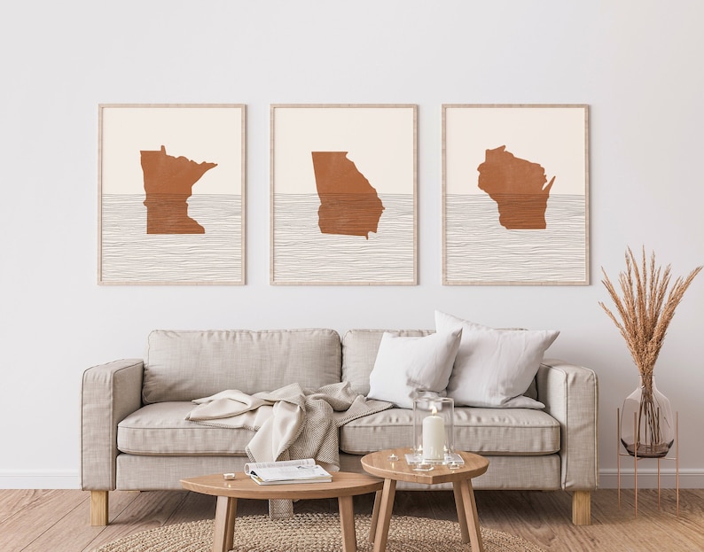 May include: Three minimalist art prints featuring the outlines of the states of Minnesota, Georgia, and Wisconsin in a burnt orange color. The states are depicted as if they are partially submerged in water, with a series of thin, horizontal lines representing the water.