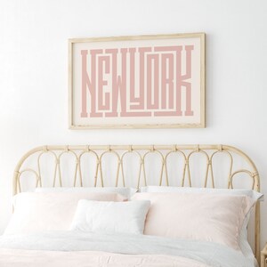 May include: A light pink framed print with the word "NEW YORK" in a bold, stylized font. The print is hanging above a light brown wicker headboard.