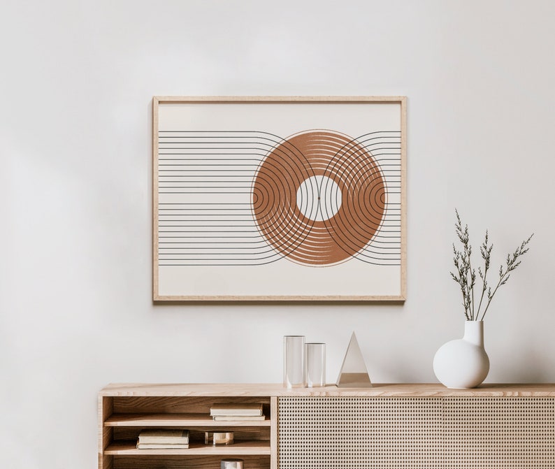 May include: A framed print with a geometric design of overlapping circles in shades of brown and white on a cream background. The print is hanging on a white wall above a wooden shelf with a white vase and dried flowers.