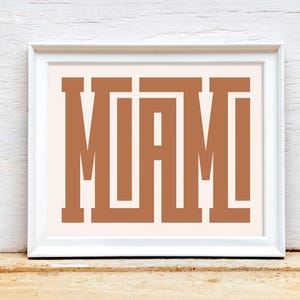 K&ouml;nnte beinhalten: Ein wei&szlig;er gerahmter Druck mit dem Wort "MIAMI" in braunen Blockbuchstaben auf einem hellrosa Hintergrund.