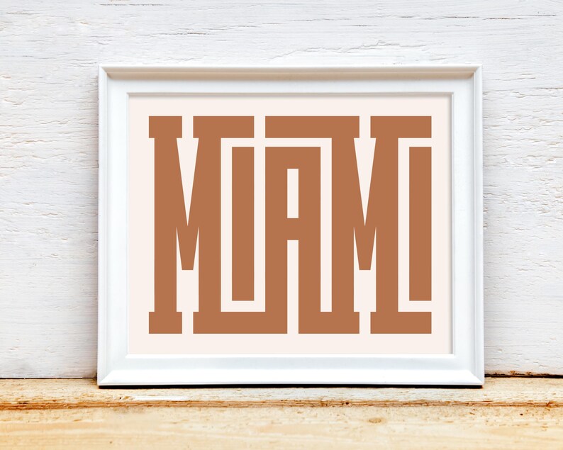 K&ouml;nnte beinhalten: Ein wei&szlig;er gerahmter Druck mit dem Wort "MIAMI" in braunen Blockbuchstaben auf einem hellrosa Hintergrund.