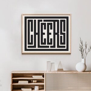 May include: A framed black and white print with the word "CHEERS" in a bold, geometric font. The print is hanging on a white wall above a wooden cabinet with a woven rattan front.