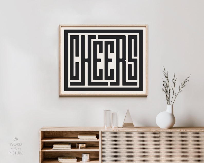 May include: A framed black and white print with the word "CHEERS" in a bold, geometric font. The print is hanging on a white wall above a wooden cabinet with a woven rattan front.