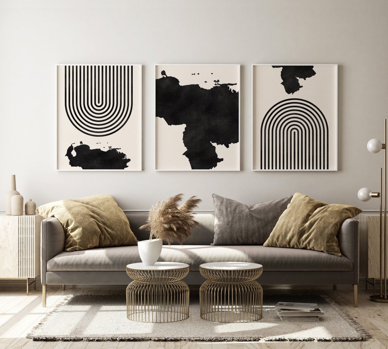 May include: A living room interior with a gray sofa, two gold metal side tables, and three black and white abstract art prints on the wall. The sofa has two brown throw pillows and a gray throw pillow. The room has a light beige wall and a light beige rug.
