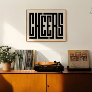 May include: A framed black and white print with the word "CHEERS" in a geometric font. The print is hanging on a white wall above a wooden cabinet with a record player and a plant.