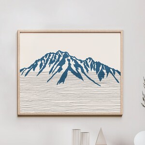 Op de afbeelding: Een minimalistische kunstprint met een blauw silhouet van een bergketen op een witte achtergrond met dunne zwarte lijnen. De print is ingelijst in een licht houten lijst.
