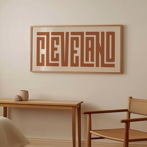 May include: A framed print with the word "CLEVELAND" in a geometric, abstract style. The print is in a light brown frame and has a white background. The word "CLEVELAND" is in a dark brown color.