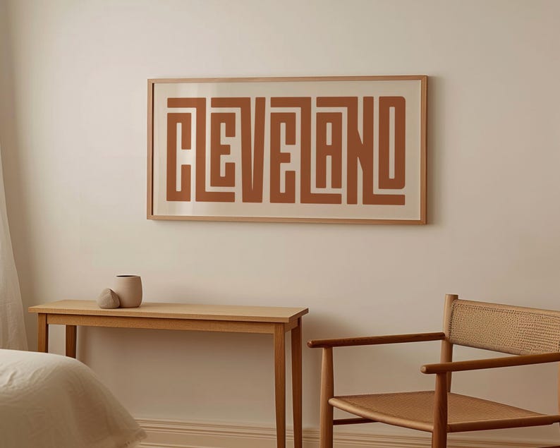 May include: A framed print with the word "CLEVELAND" in a geometric, abstract style. The print is in a light brown frame and has a white background. The word "CLEVELAND" is in a dark brown color.