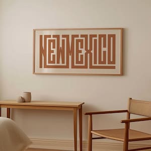 May include: A framed print on a wall features the word "NEW MEXICO" in a geometric, maze-like design. The print is in shades of brown and beige. A wooden table and chair are in front of the wall, with a small vase on the table.