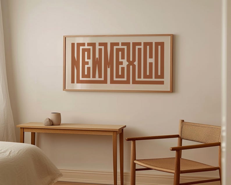 May include: A framed print on a wall features the word "NEW MEXICO" in a geometric, maze-like design. The print is in shades of brown and beige. A wooden table and chair are in front of the wall, with a small vase on the table.