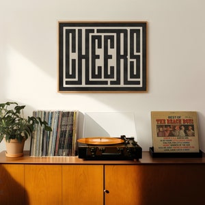 May include: A black and white framed print with the word "CHEERS" in a geometric design. The print is hanging on a white wall above a wooden cabinet with a record player, a stack of vinyl records, and a potted plant.