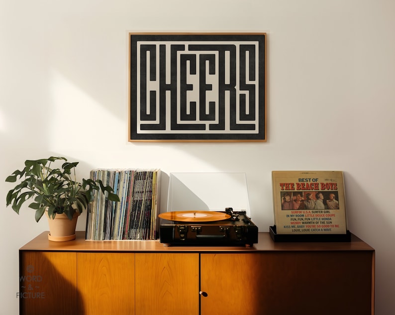 May include: A black and white framed print with the word "CHEERS" in a geometric design. The print is hanging on a white wall above a wooden cabinet with a record player, a stack of vinyl records, and a potted plant.