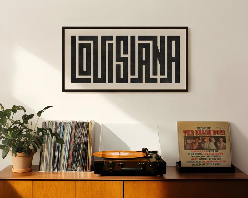 Puede incluir: Una impresi&oacute;n enmarcada en blanco y negro con la palabra "LOUISIANA" en una fuente geom&eacute;trica audaz. La impresi&oacute;n est&aacute; colgada en una pared blanca sobre un tocadiscos y una estanter&iacute;a de discos de vinilo.