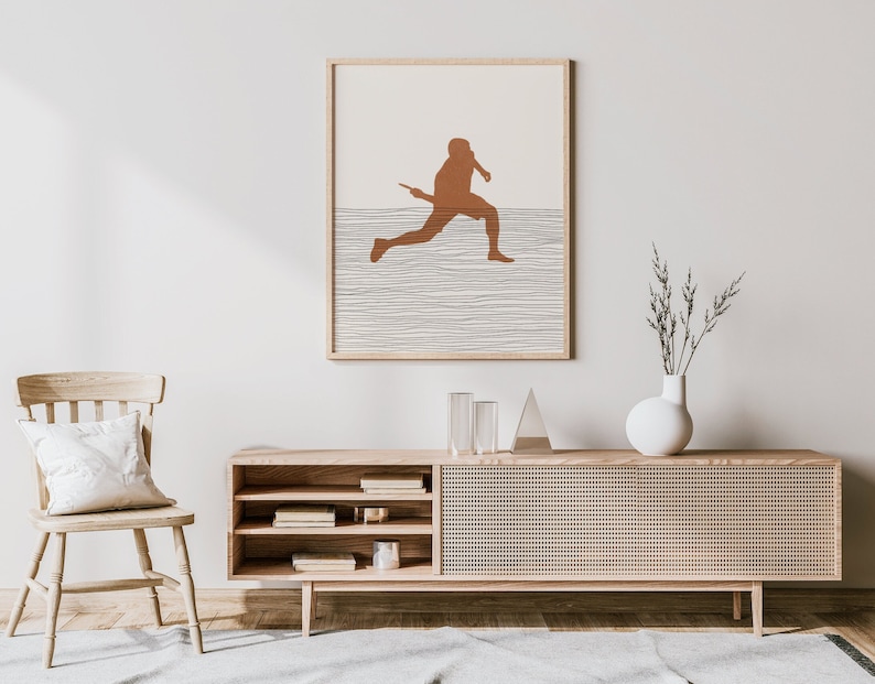 May include: A framed art print featuring a minimalist design of a baseball player in a running pose. The artwork uses a warm brown color for the figure against a neutral background with horizontal lines. The print is displayed above a wooden cabinet.
