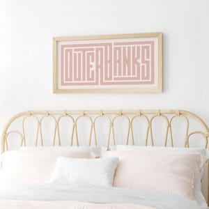 May include: A pink and white framed print with the text "OUTER BANKS" in a geometric font. The print is hanging above a light brown wicker headboard with a white and pink bedding set.