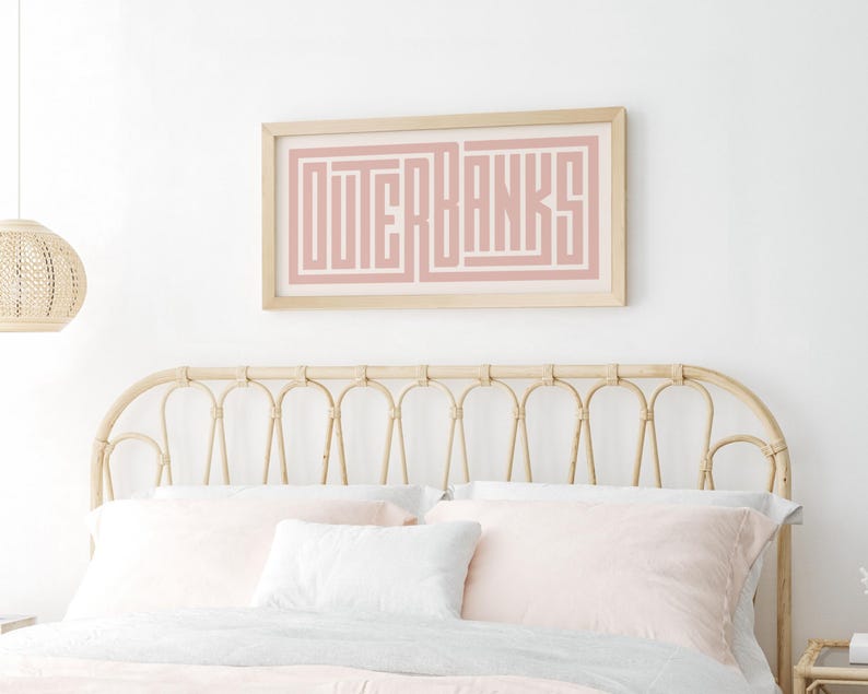 May include: A pink and white framed print with the text "OUTER BANKS" in a geometric font. The print is hanging above a light brown wicker headboard with a white and pink bedding set.