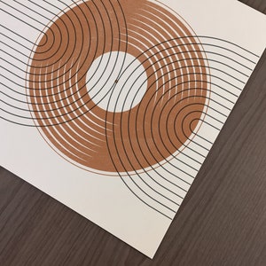 May include: Abstract art print with a brown circle design on a white background. The circle is made up of concentric rings with black lines. The background is a series of black lines.