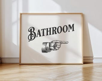 以下が含まれることがあります： 装飾的なフォントで「BATHROOM」という言葉が書かれた額入りのプリント。右を指す手も描かれています。アートワークは白黒で、ヴィンテージ感があります。フレームは明るい木の色です。
