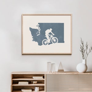 May include: A framed print of a blue silhouette of the state of Washington with a mountain biker riding through the state. The print is on a white background and is framed in a light wood frame.
