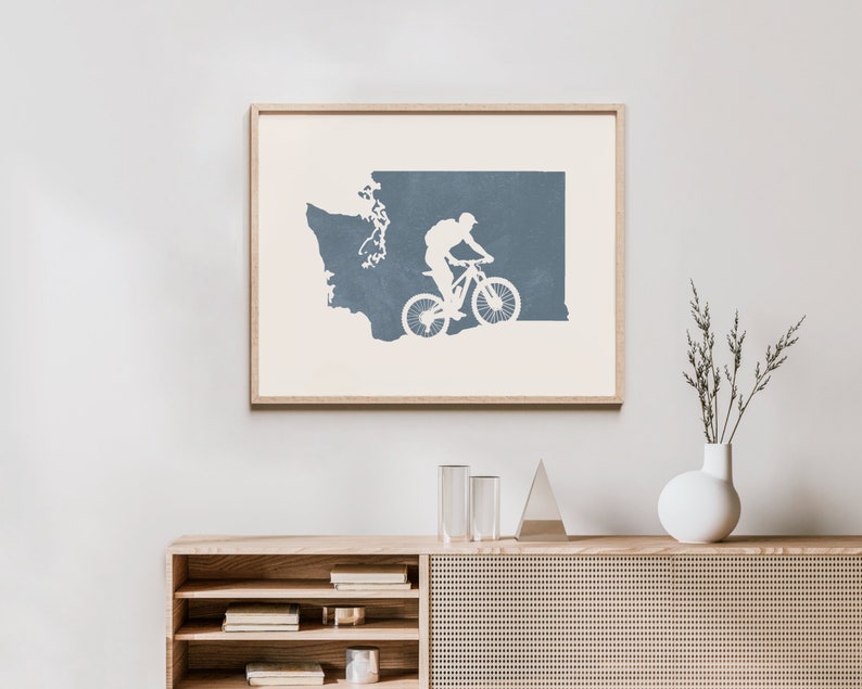 May include: A framed print of a blue silhouette of the state of Washington with a mountain biker riding through the state. The print is on a white background and is framed in a light wood frame.
